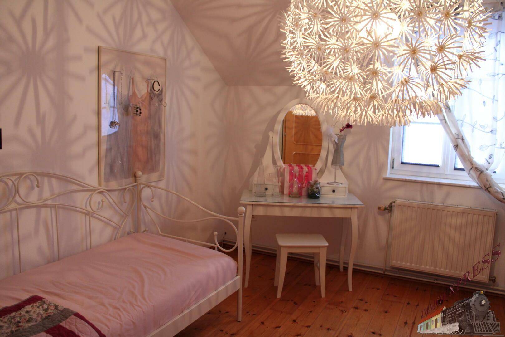 Mädchenzimmer mit weißem Metallbett, Schminktisch und auffälliger Deckenleuchte, sehr hell.