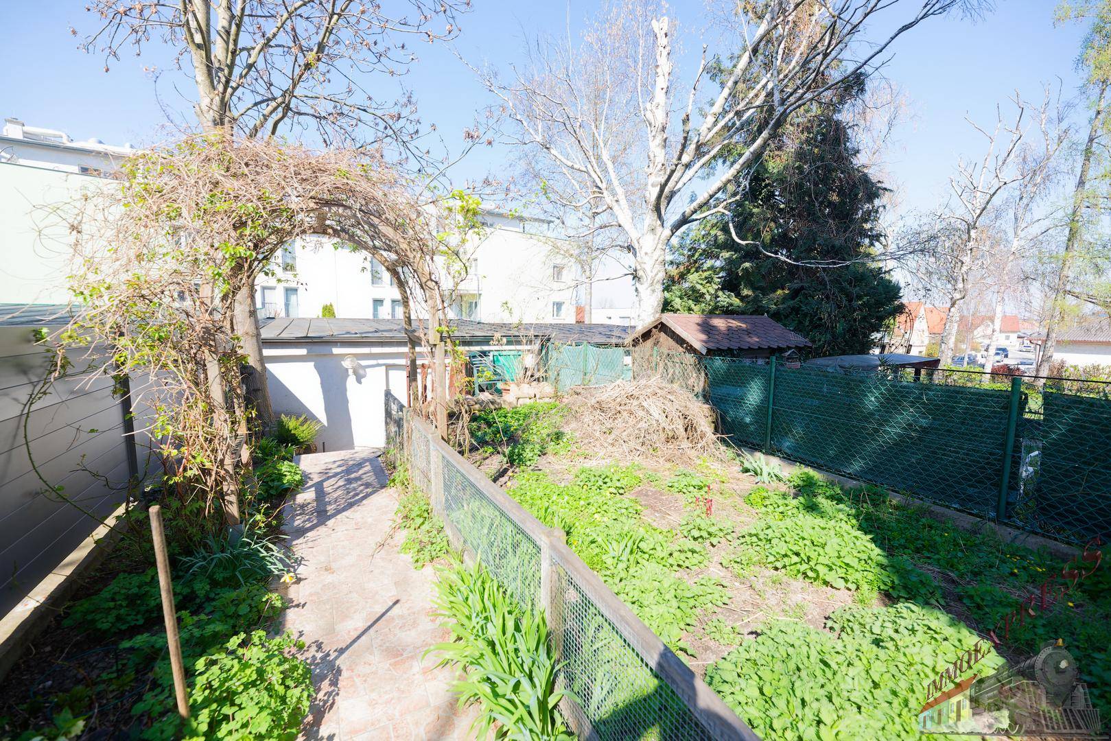 Gartenbereich mit Gartenhaus, Zaun und bewachsenem Torbogen.