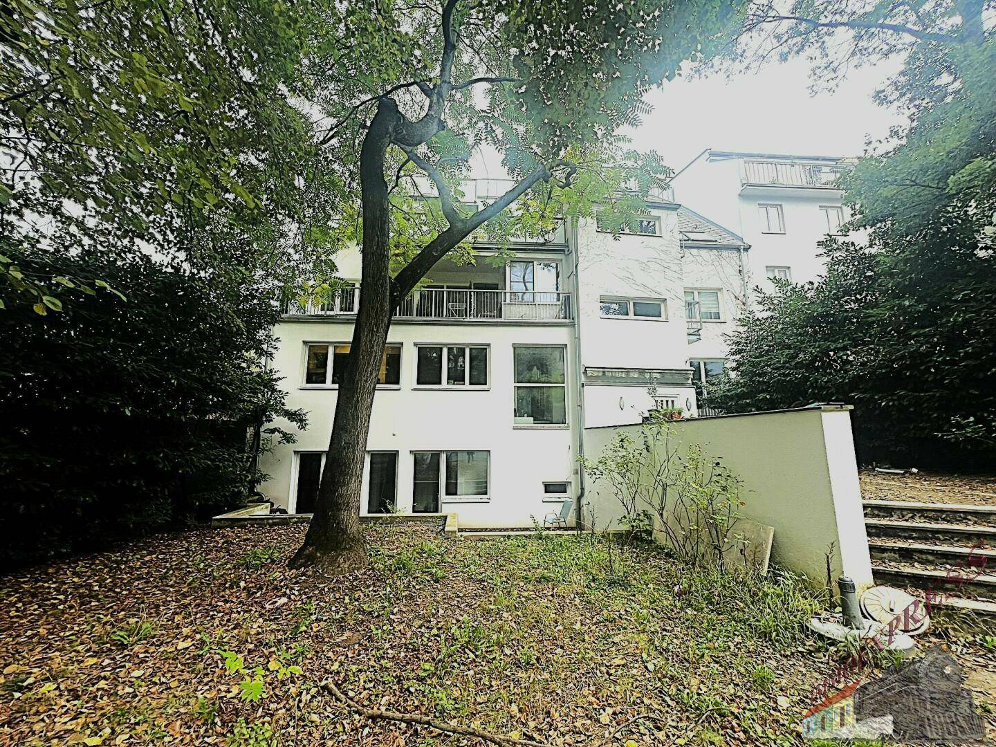 Rückansicht eines Mehrfamilienhauses mit mehreren Balkonen, umgeben von Bäumen und einem Gartenbereich.