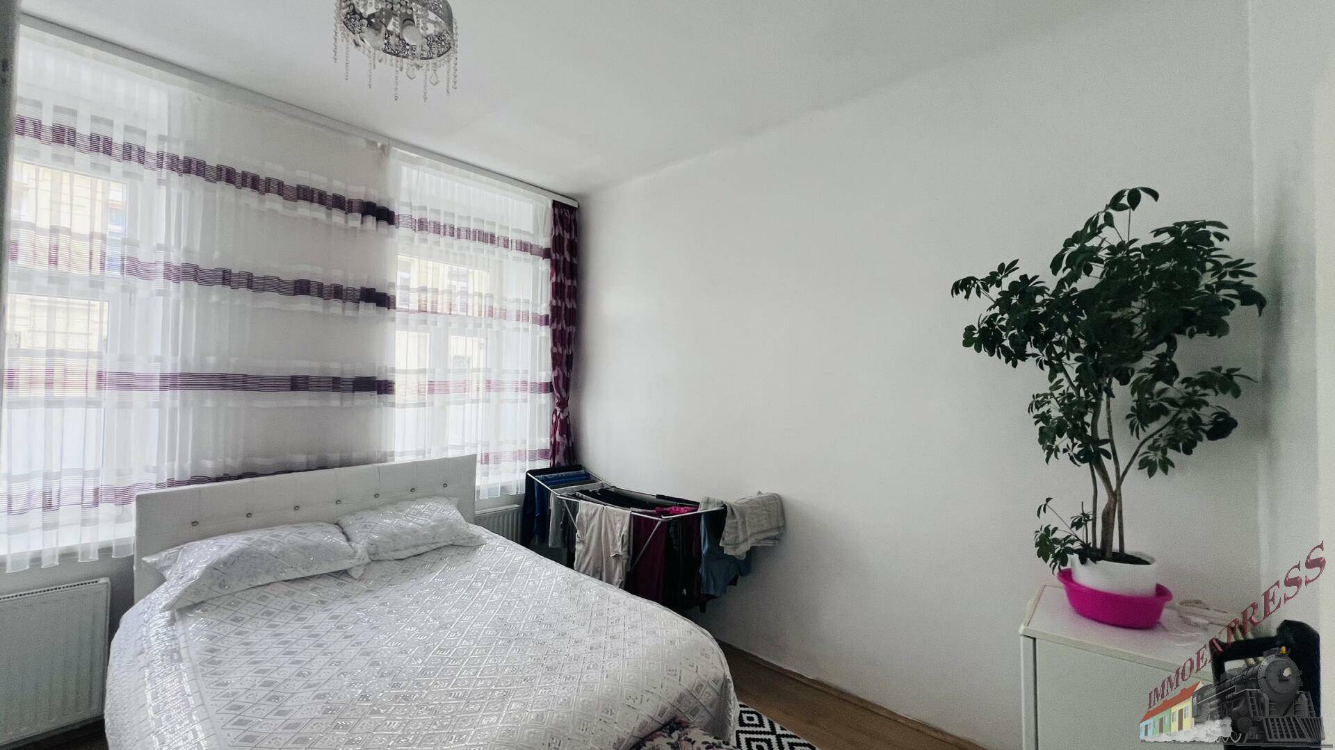 Helles Schlafzimmer mit Doppelbett, Wäscheständer und großem Fenster, das viel Licht hereinlässt.