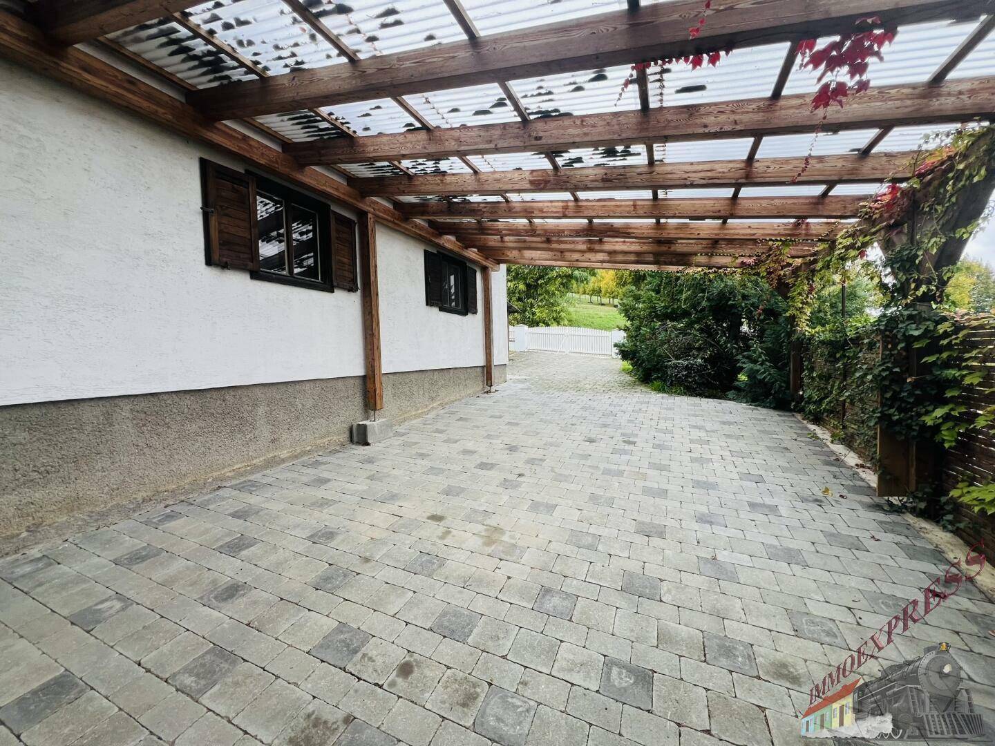 Überdachter Stellplatz mit Holzpergola und gepflastertem Boden neben dem Haus.