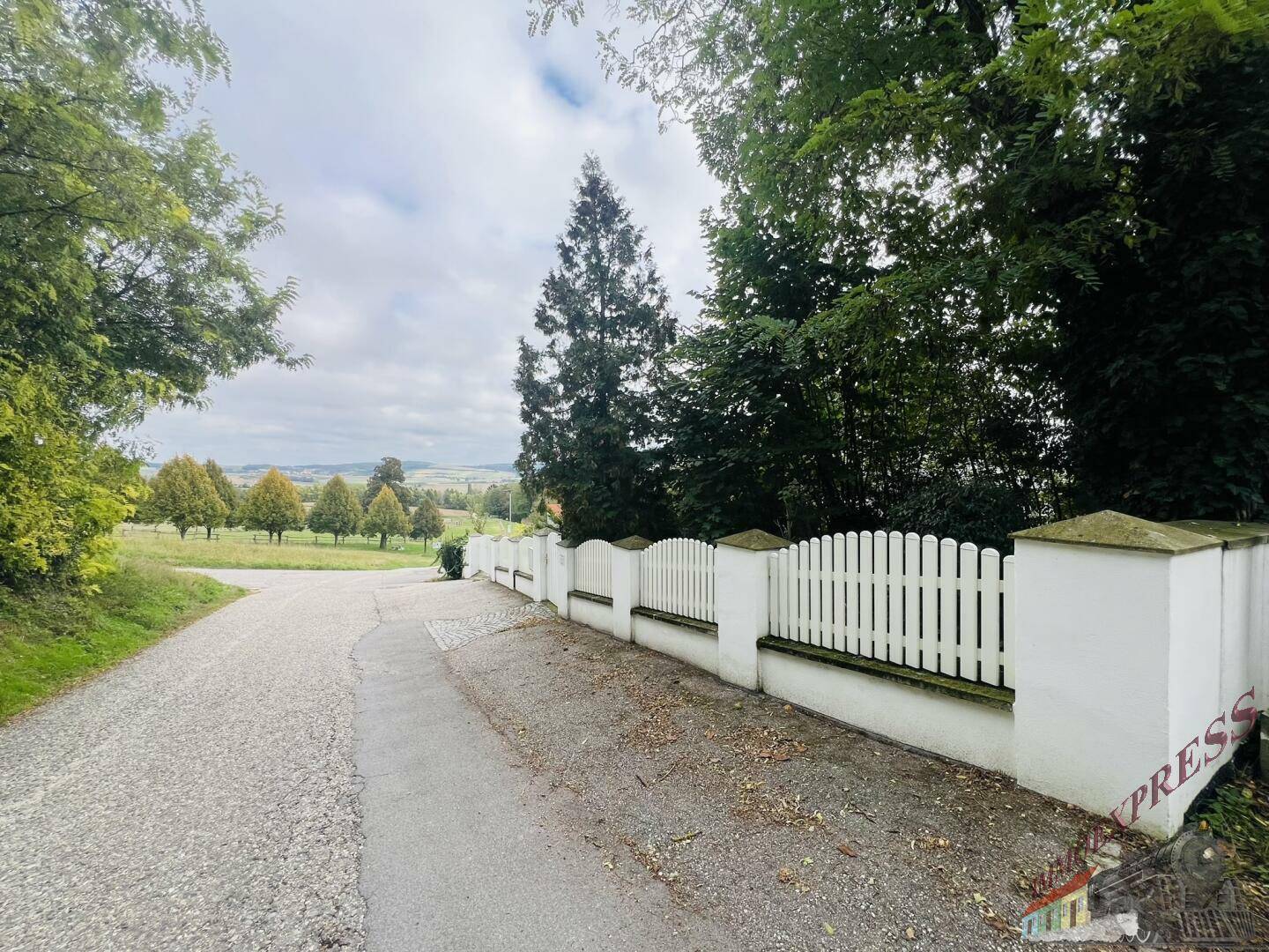 Landstraße mit weißem Zaun und weitem Blick über die grüne, hügelige Landschaft.