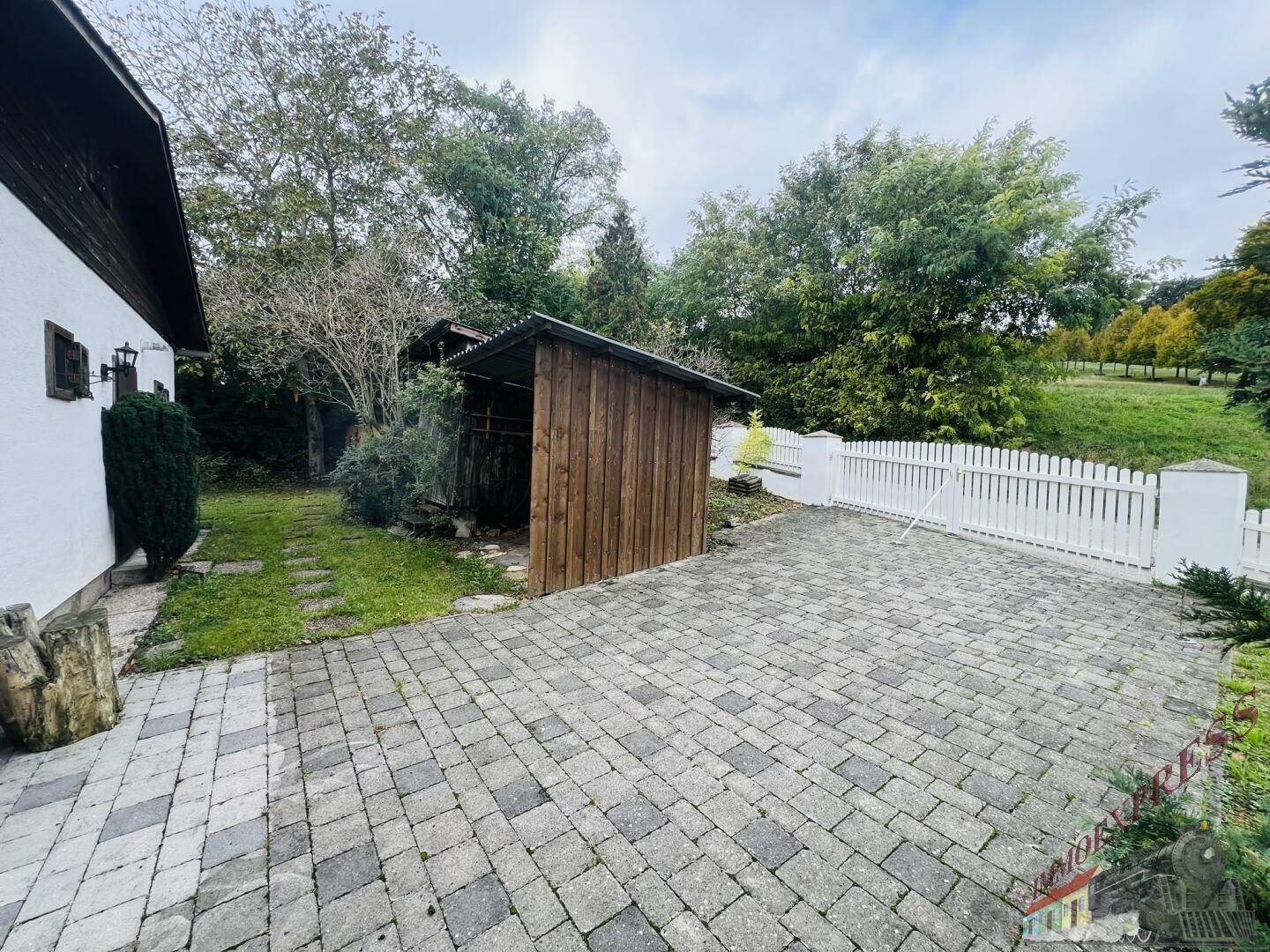 Gepflasterter Bereich mit Holzschuppen und weißem Zaun, umgeben von Grün.