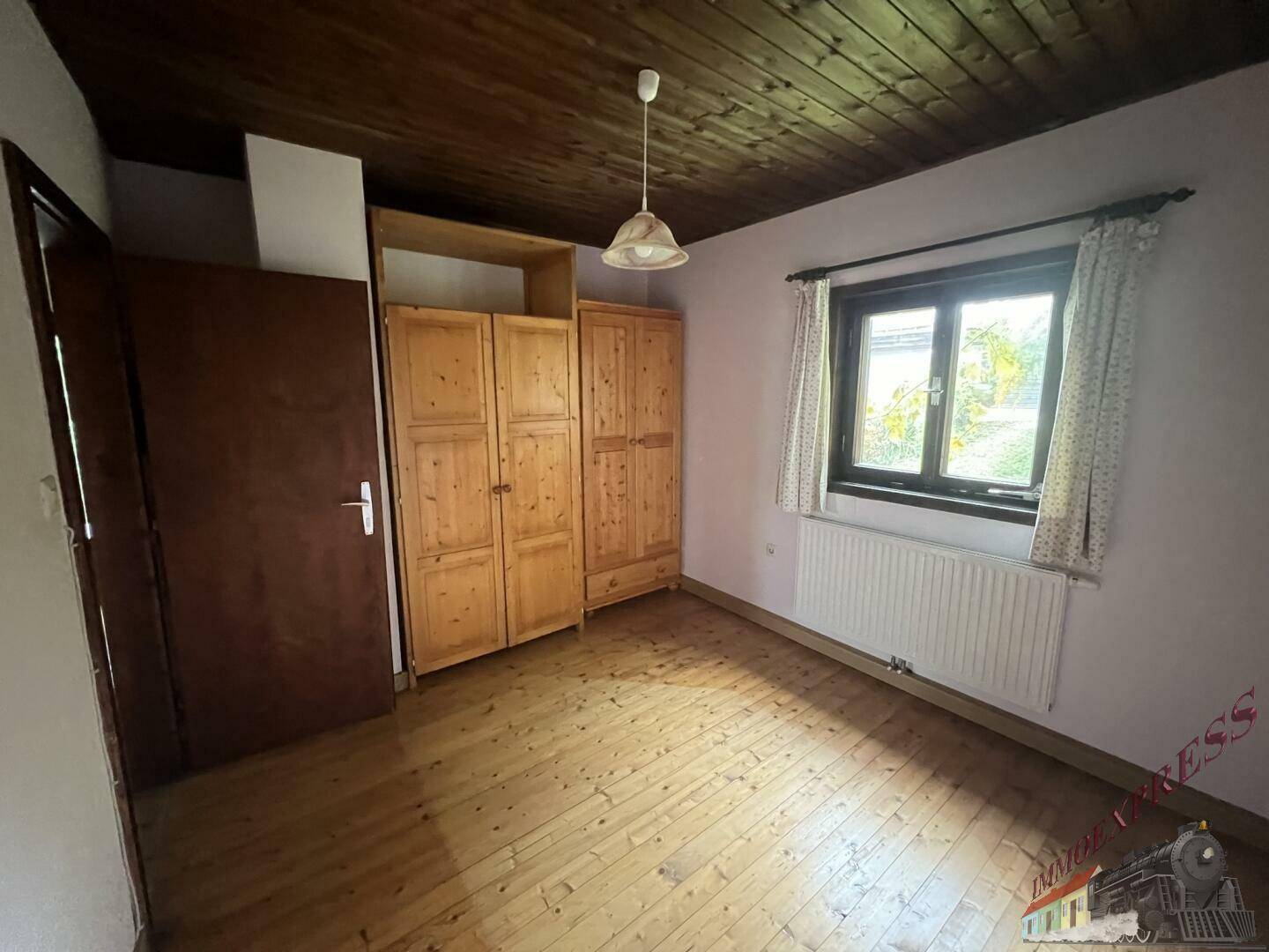 Schlafzimmer mit Holzdielenboden, Holzdecke und einem Holzschrank neben dem Fenster.