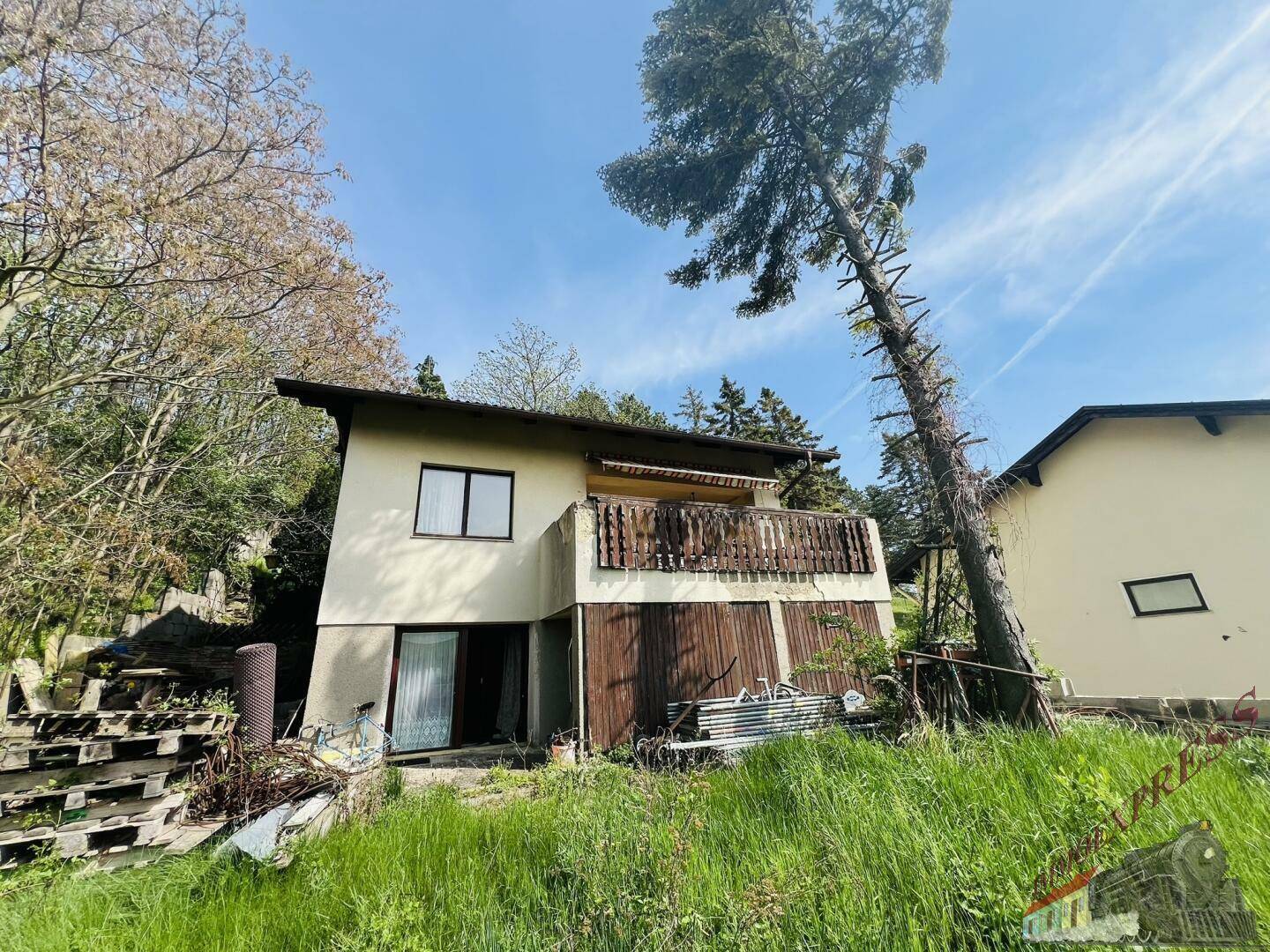 Älteres Haus mit Balkon und bewachsenem Hang, umgeben von Bäumen.
