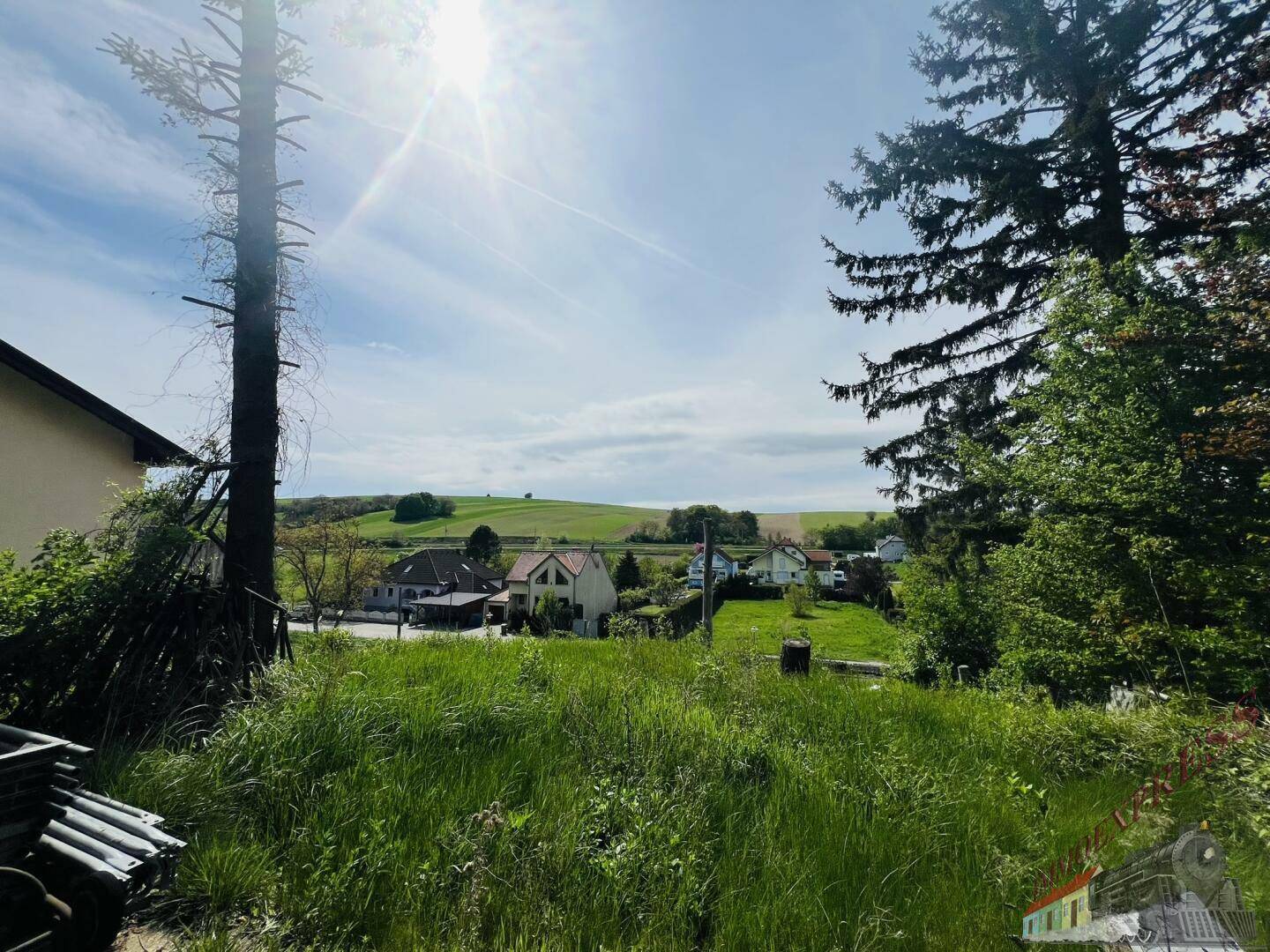 Sonnige Landschaft mit weiten Feldern und vereinzelten Häusern unter blauem Himmel.