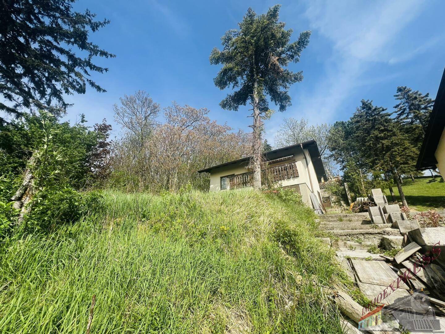 Haus auf einem bewachsenen Hang mit Bäumen im Hintergrund unter blauem Himmel.