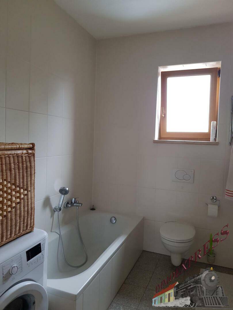 Badezimmer mit Badewanne, Toilette und Fenster, ausgestattet mit weißen Fliesen.
