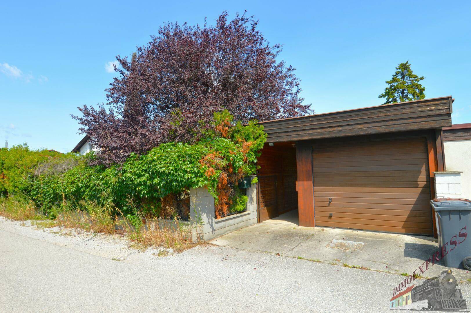 Garage mit Holztor und Zufahrt, umgeben von üppiger Vegetation.