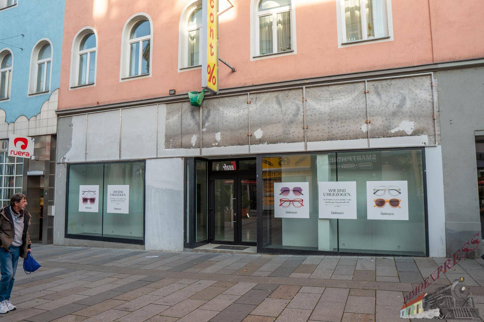 Fassade eines älteren Geschäftsgebäudes mit großen Schaufenstern und einem renovierungsbedürftigen oberen Bereich.