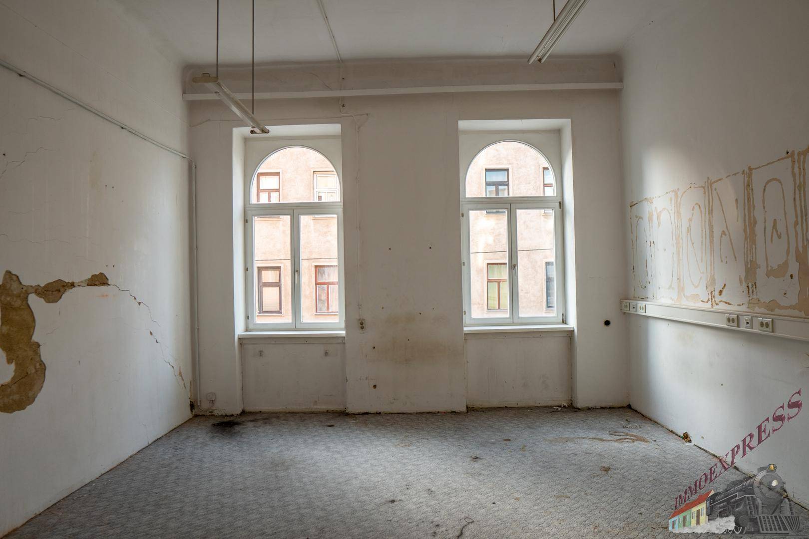 Unrenovierter Raum mit Rundbogenfenstern, Teppichboden und deutlichen Abnutzungsspuren an den Wänden.
