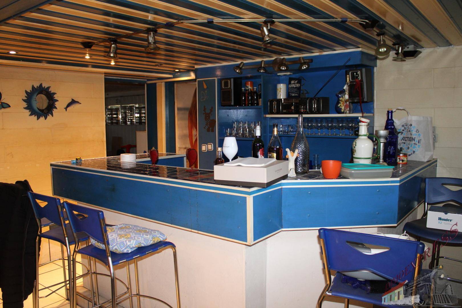 Kellerbar mit blauer Holzverkleidung, Barhockern und Beleuchtung.