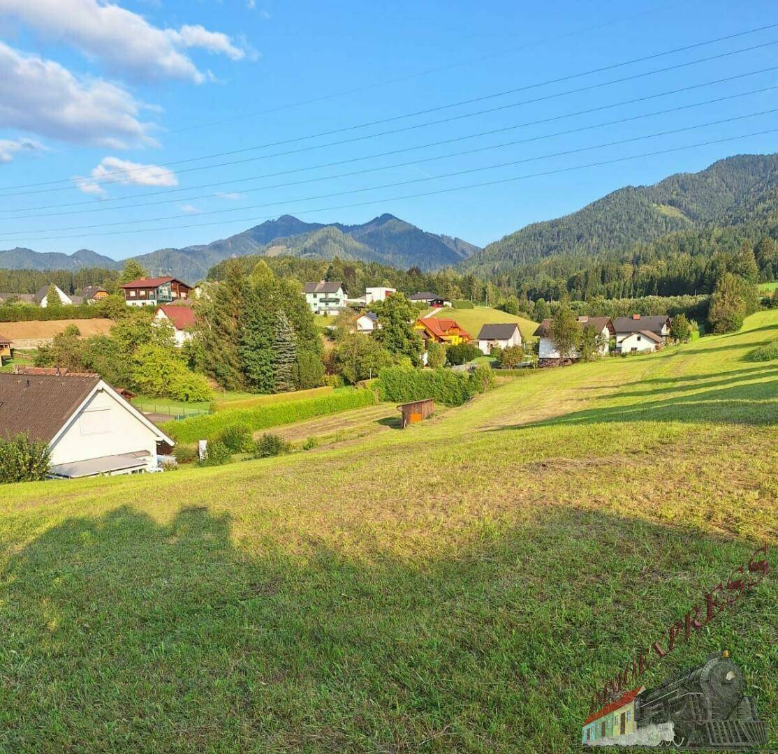 Idyllisches Grundstück mit grüner Wiese und weitem Blick auf die sonnige Berglandschaft und Häuser.