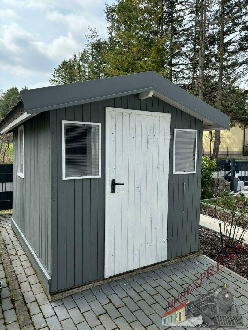 Kleines, graues Gartenhaus mit weißer Holztür und Fenstern auf gepflastertem Untergrund.
