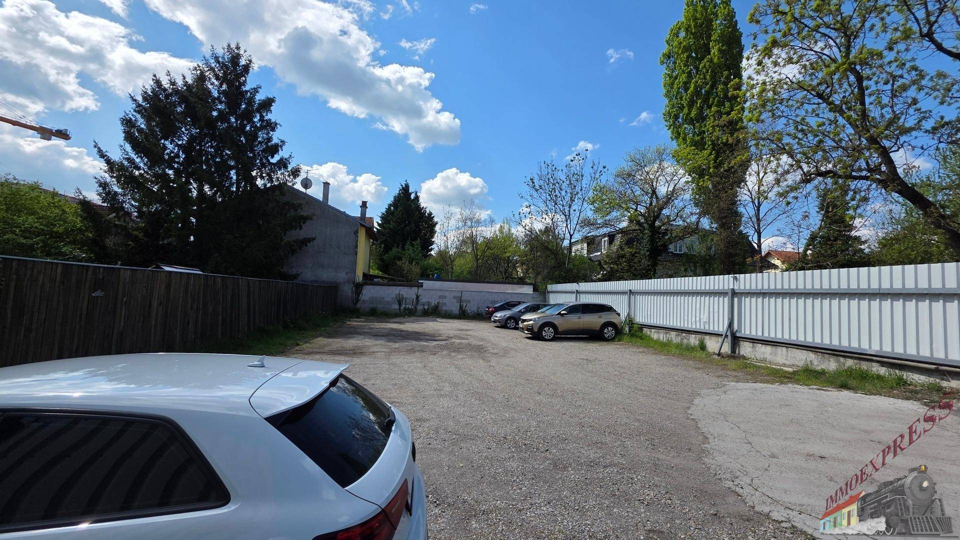 Großer Schotterparkplatz mit mehreren Autos, umgeben von Zäunen und Bäumen unter blauem Himmel.