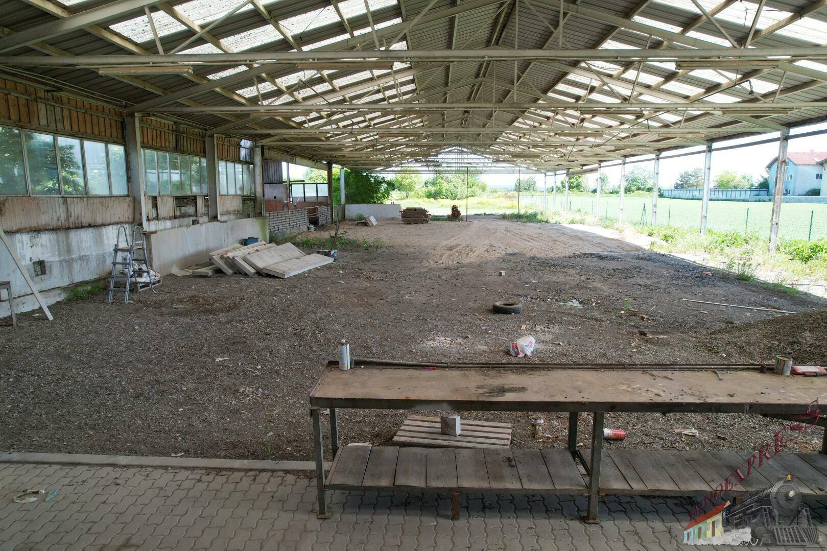 Innenansicht einer großen, offenen Halle mit Erdboden, sichtbarer Dachkonstruktion und einer Werkbank.