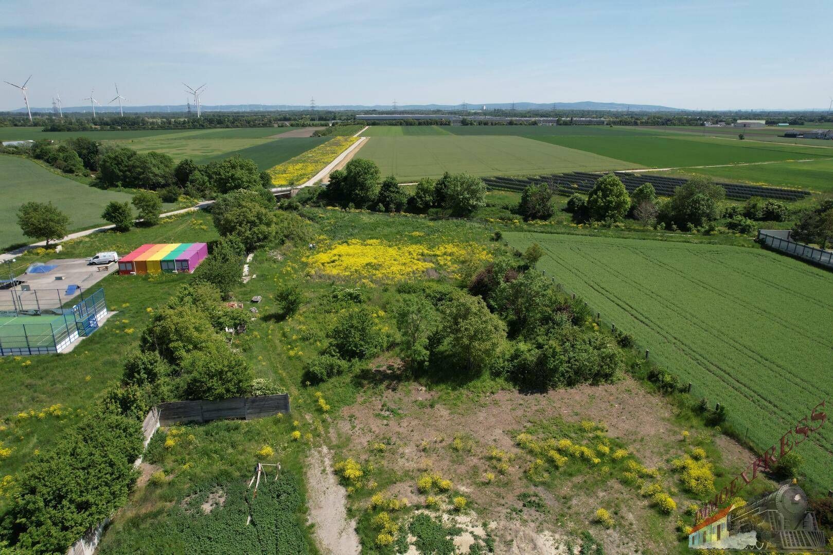 Weitläufige Luftaufnahme eines ländlichen Gebiets mit Feldern, Gebäuden und Windrädern am Horizont.