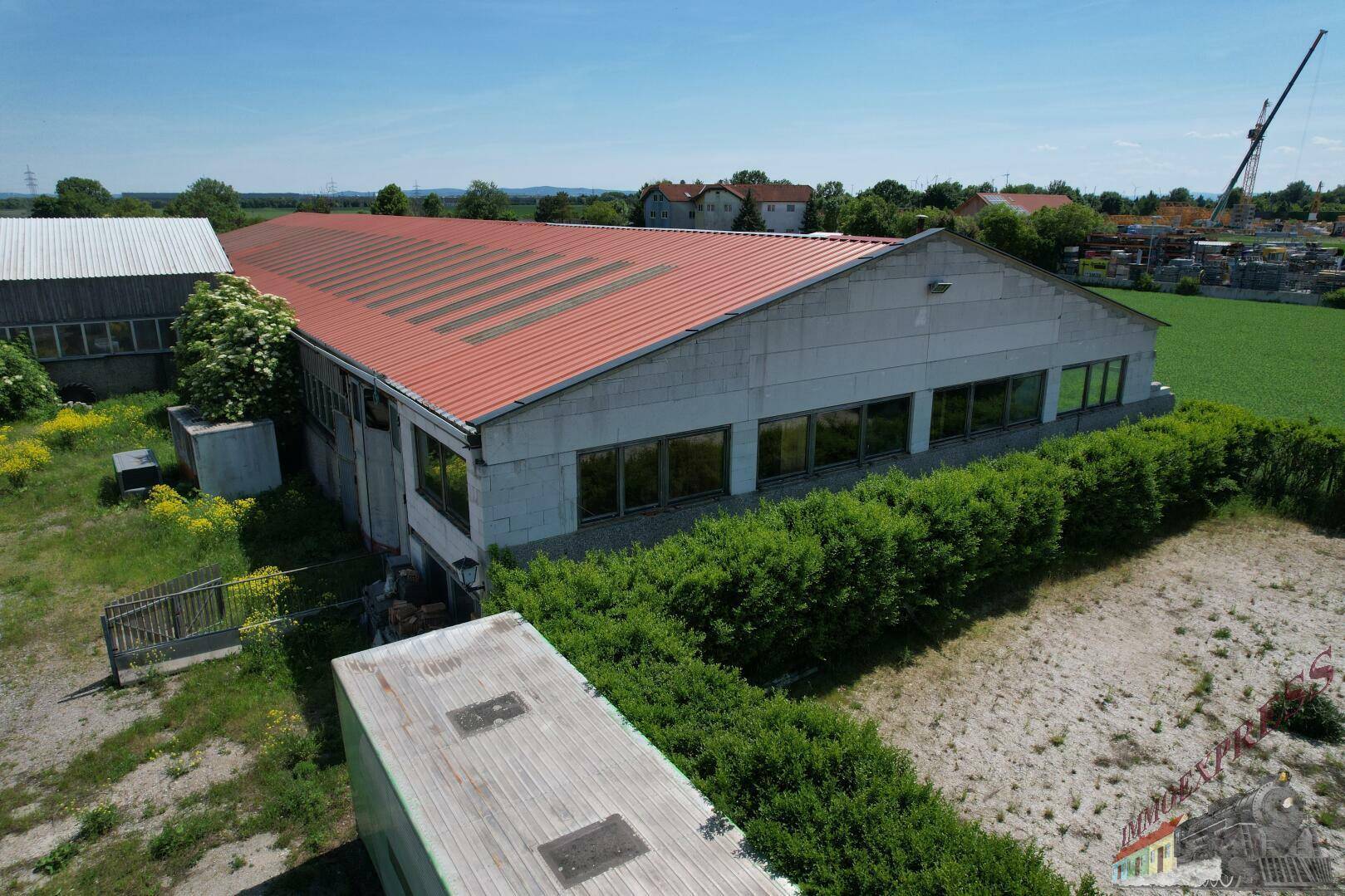 Luftaufnahme einer langen Halle mit rotem Dach und großen Fensterfronten, umgeben von einer Hecke und Feldern.