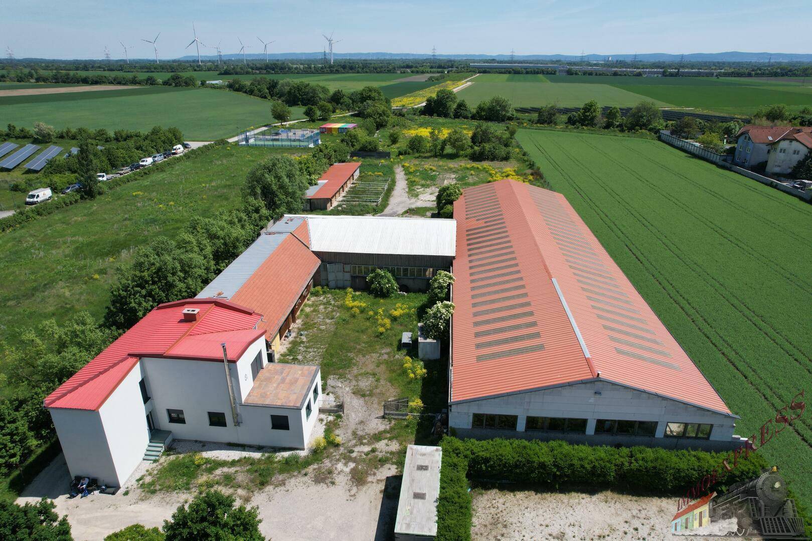 Umfassende Luftaufnahme des gesamten Anwesens mit mehreren Gebäuden, Feldern und Windrädern in der Ferne.