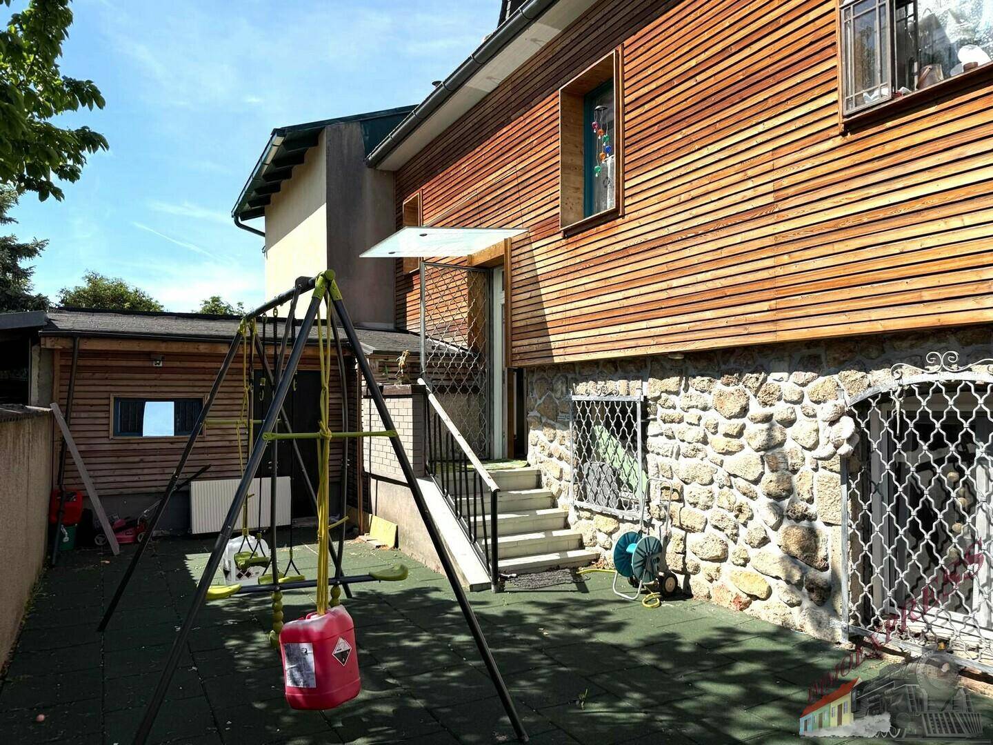 Hinterhofbereich des Hauses mit Kinderspielplatz, Schaukel und rustikaler Stein- und Holzfassade.