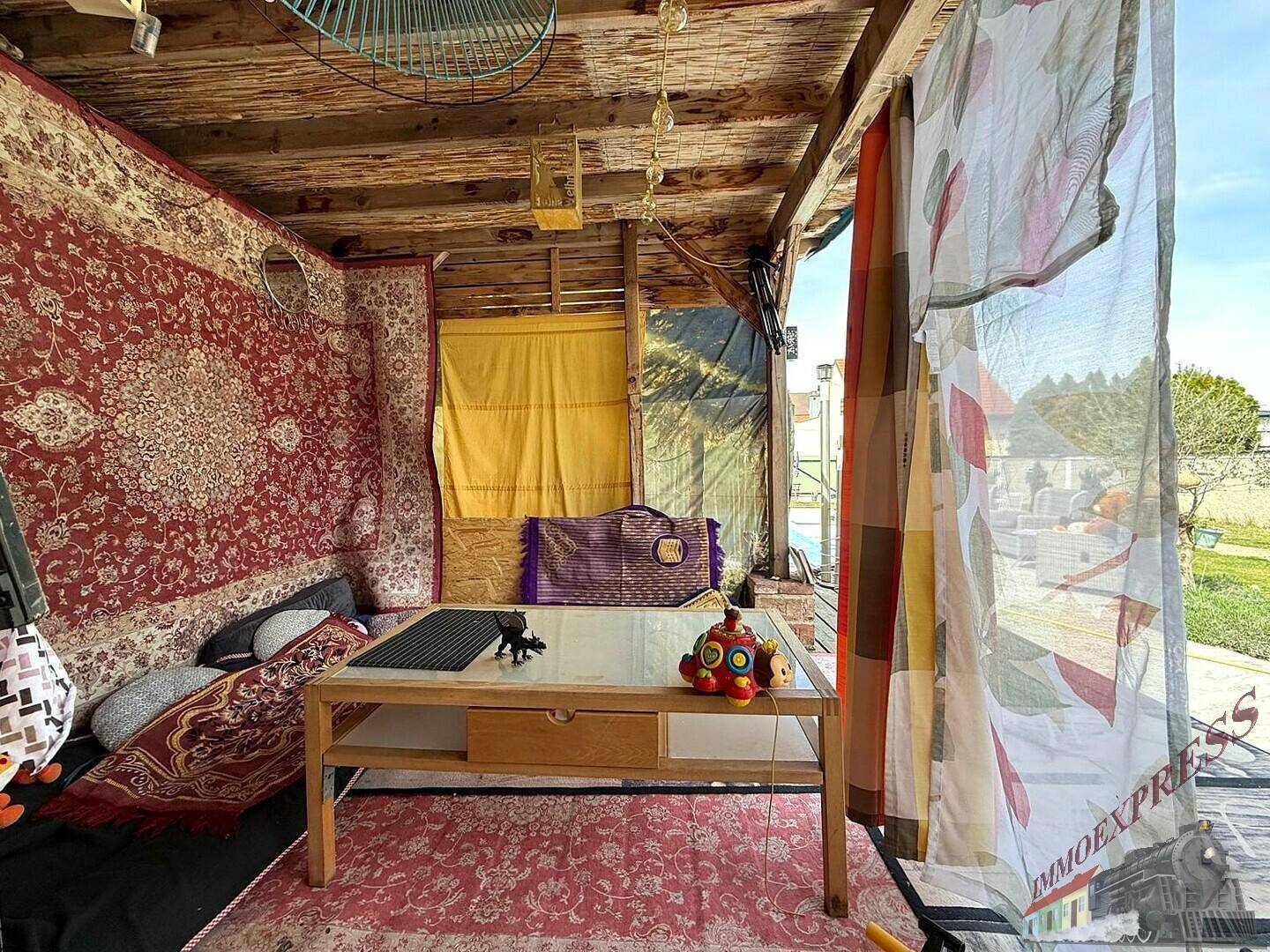Gemütlicher, überdachter Außenwohnbereich mit Teppichen, Holztisch und farbenfroher Dekoration.