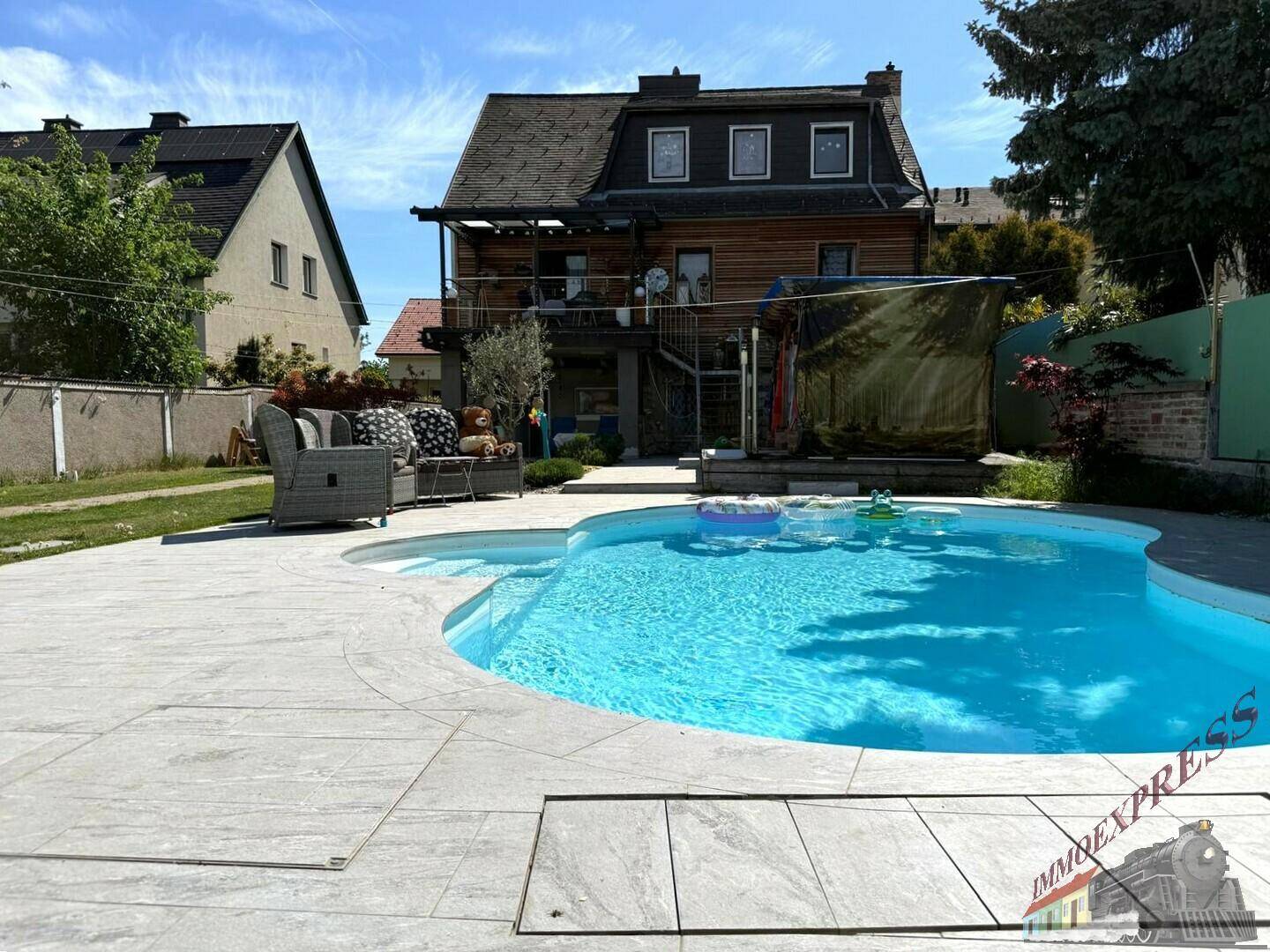 Hinteransicht des Hauses mit großem Pool, Terrasse und Gartenmöbeln unter blauem Himmel.