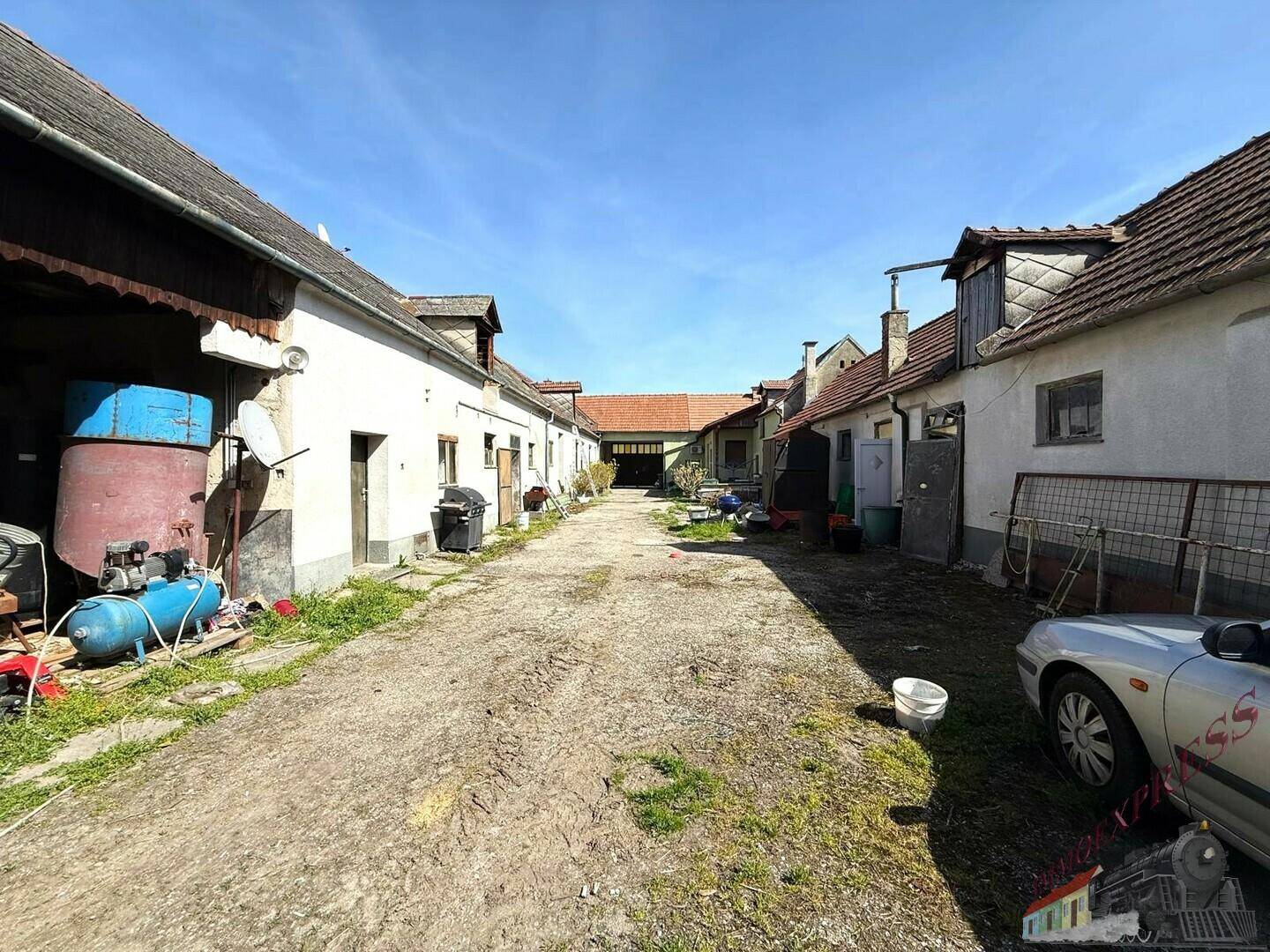Langer, unbefestigter Innenhof zwischen mehreren alten landwirtschaftlichen Gebäuden mit verschiedenen Gegenständen.