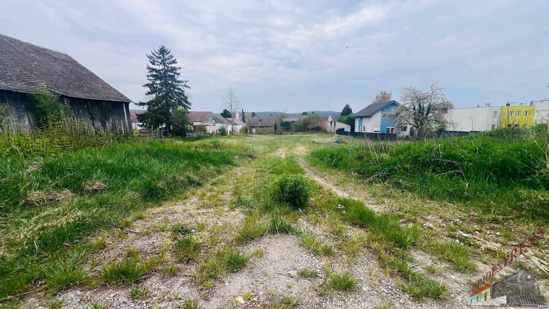 Unbebautes Grundstück mit Schotterweg, umgeben von grüner Landschaft und vereinzelten Gebäuden.