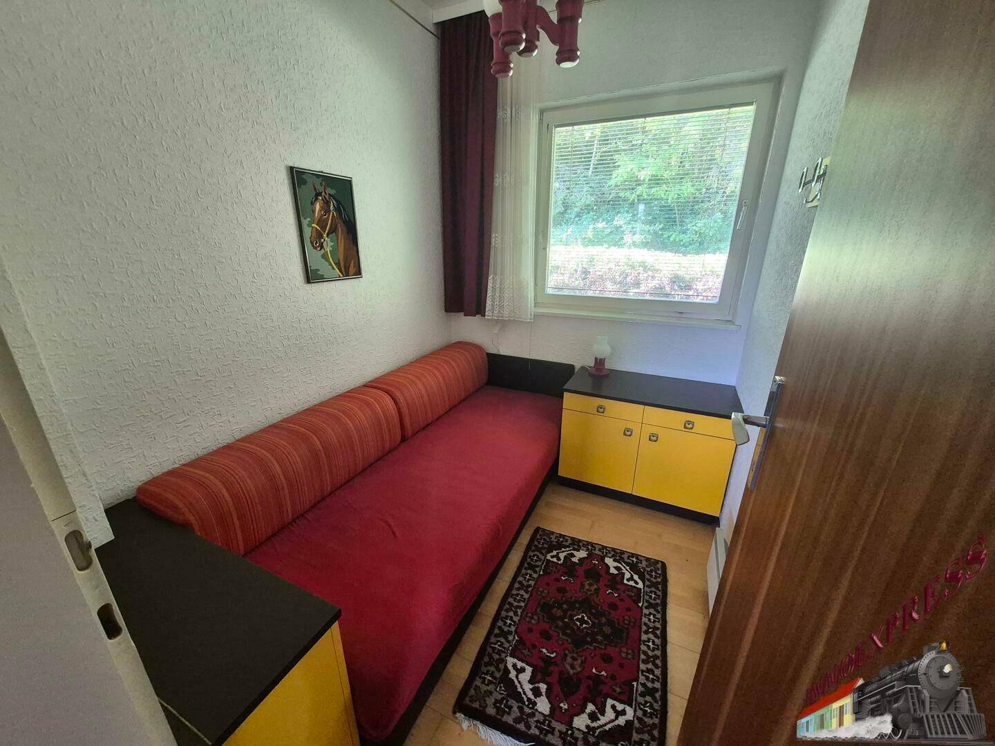 Kleines Schlafzimmer mit rotem Sofa, Holzfußboden und Fenster.