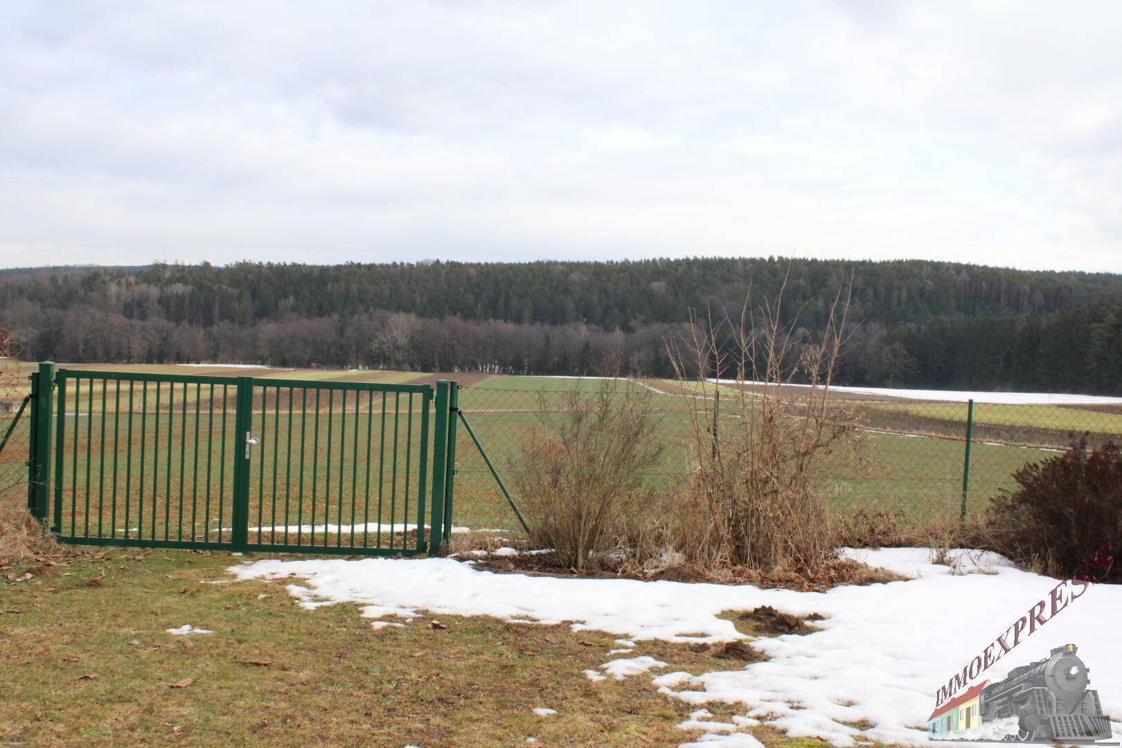 Blick über Felder und Wald mit einem grünen Zaun im Vordergrund.