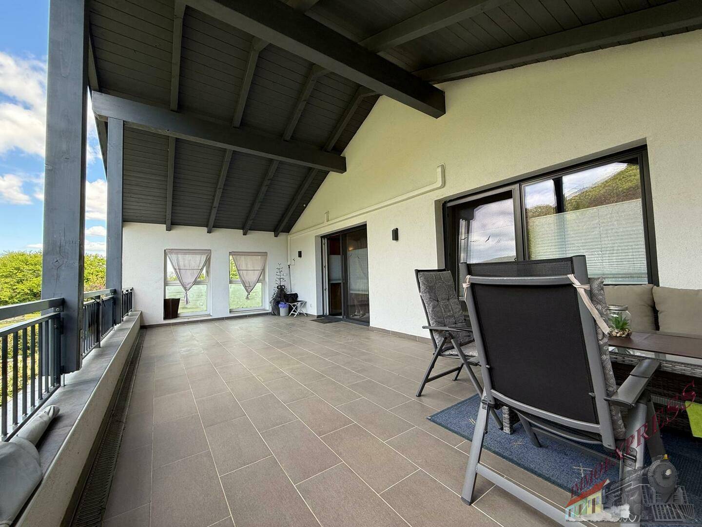 Großer überdachter Balkon mit Fliesenboden, Sitzgelegenheiten und weitem Blick ins Grüne.