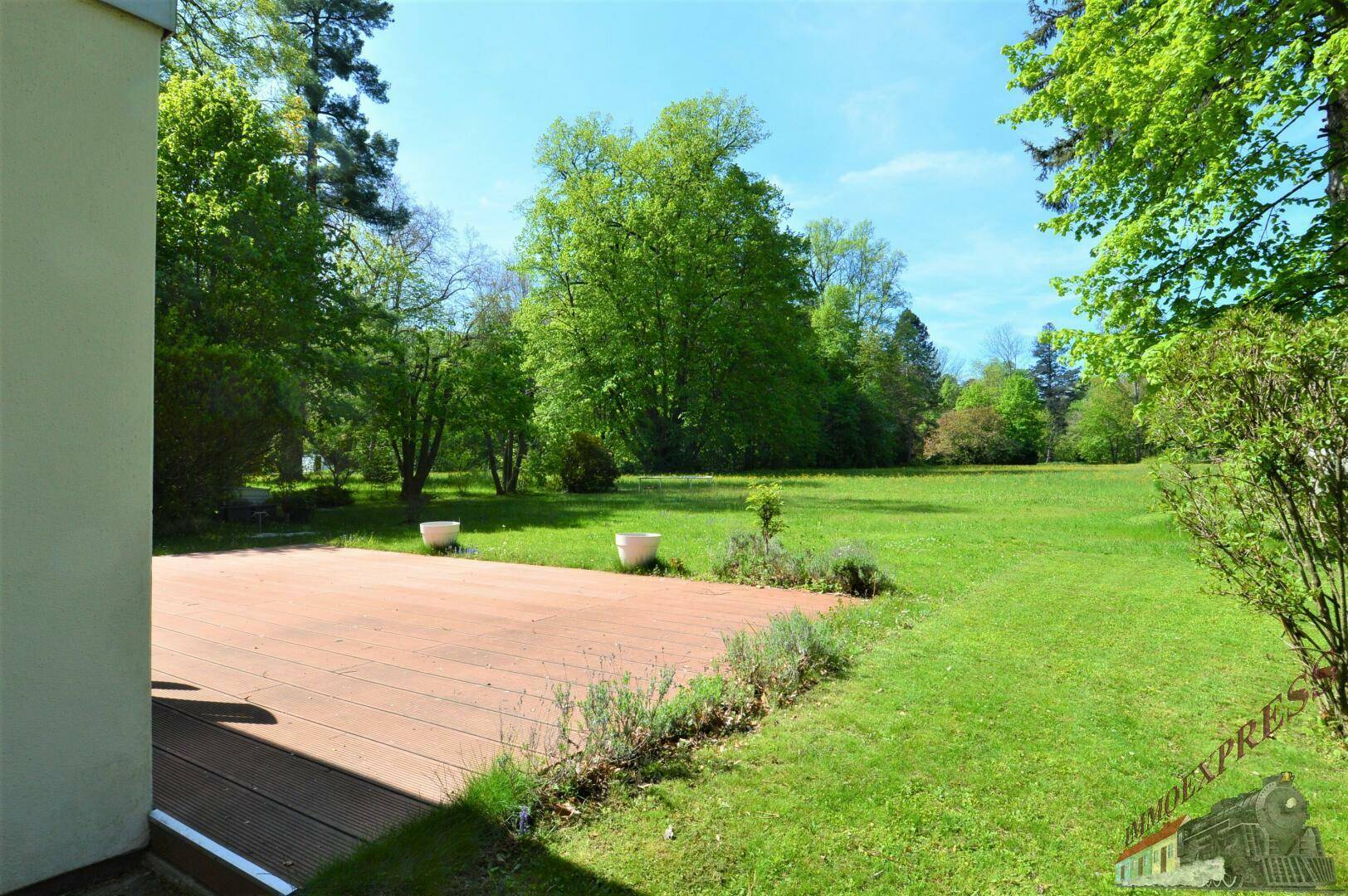 Eine Holzterrasse grenzt an eine weitläufige grüne Rasenfläche mit Bäumen im Hintergrund.