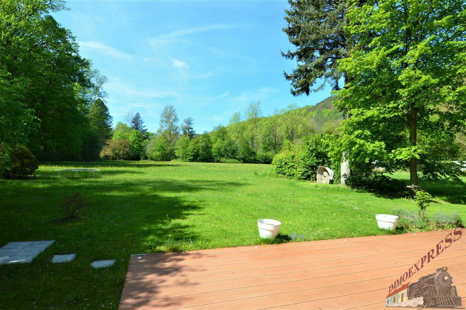 Eine weitläufige grüne Rasenfläche mit einer Holzterrasse im Vordergrund und bewaldeten Hügeln im Hintergrund.