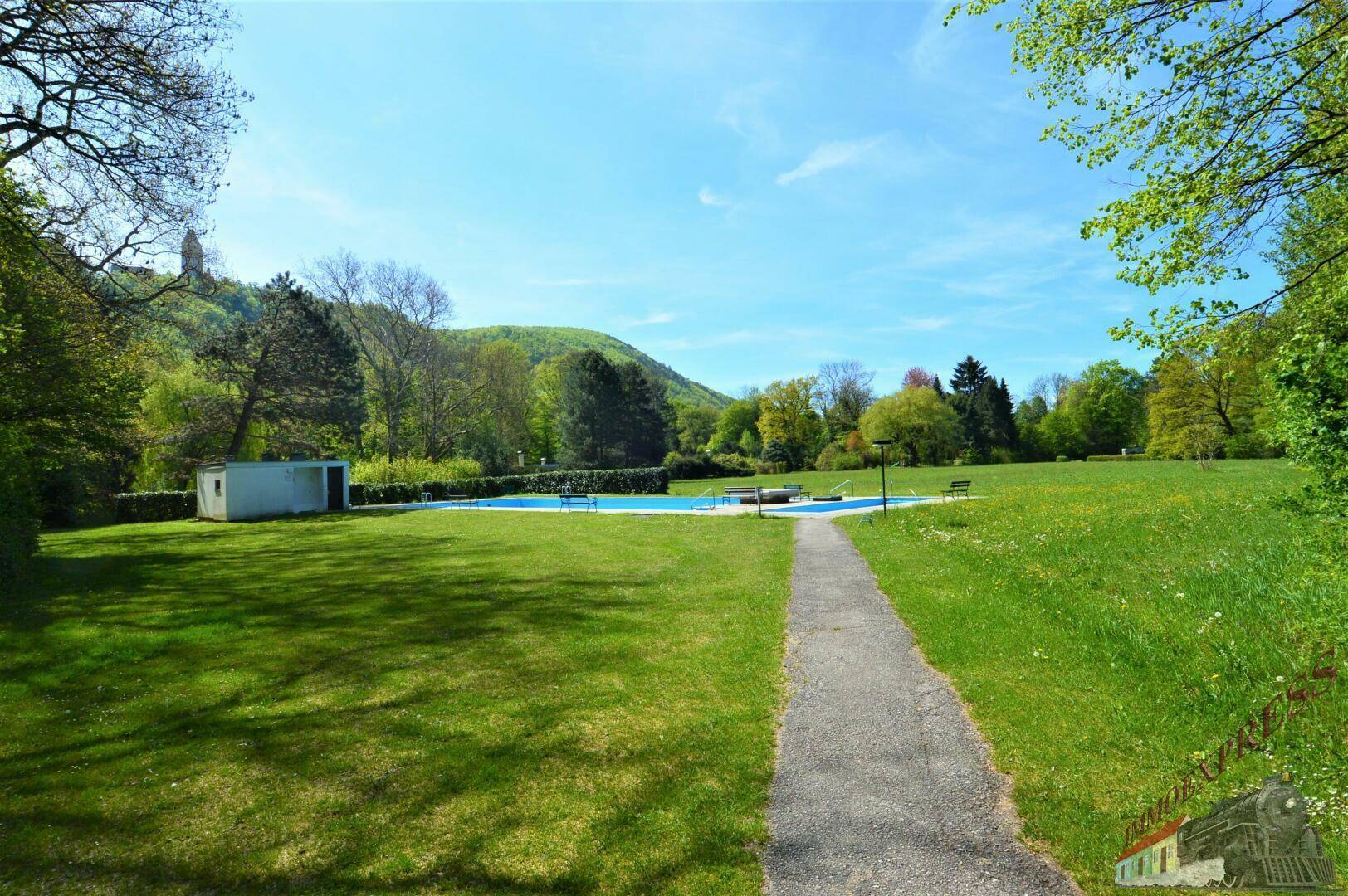 Panoramablick über eine weite grüne Wiese mit einem Pool und bewaldeten Hügeln im Hintergrund.