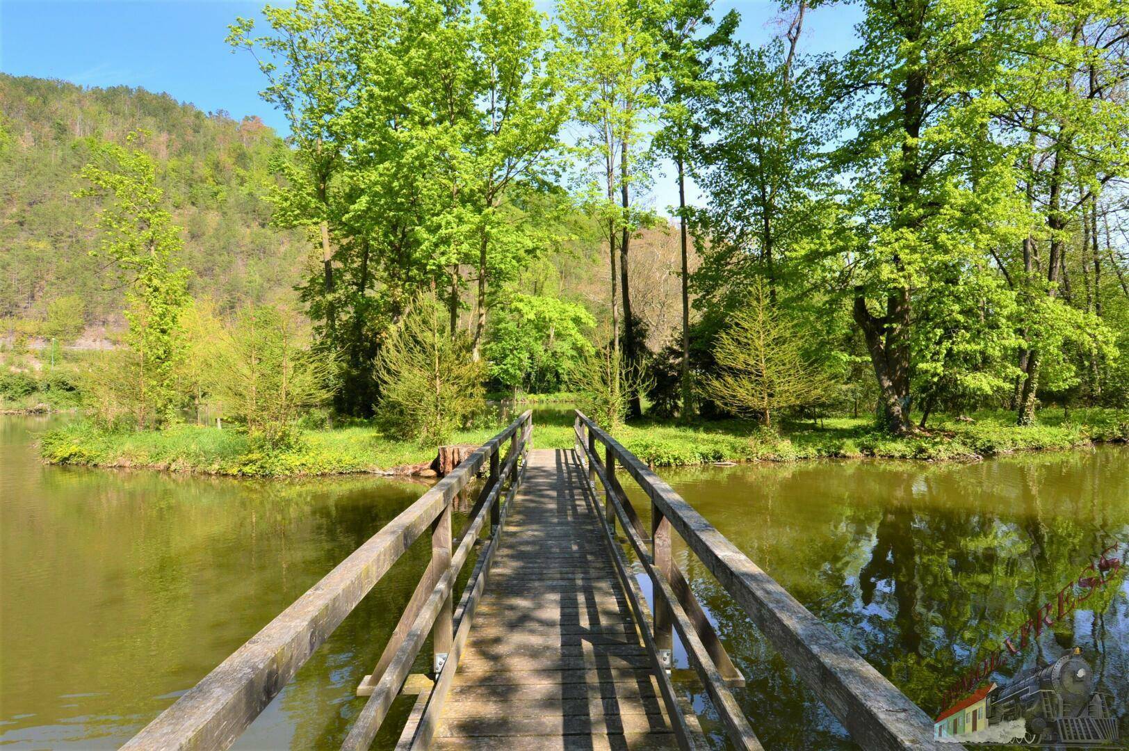 Hölzerne Brücke über einen Teich, umgeben von üppiger grüner Natur und bewaldeten Hügeln.