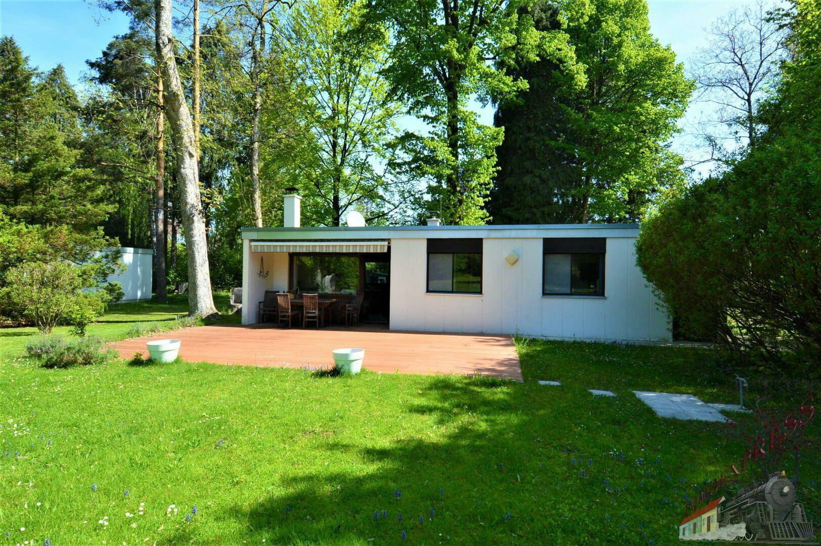 Ansicht des Hauses von hinten mit Terrasse, Gartenmöbeln und dem umgebenden grünen Rasen und Bäumen.