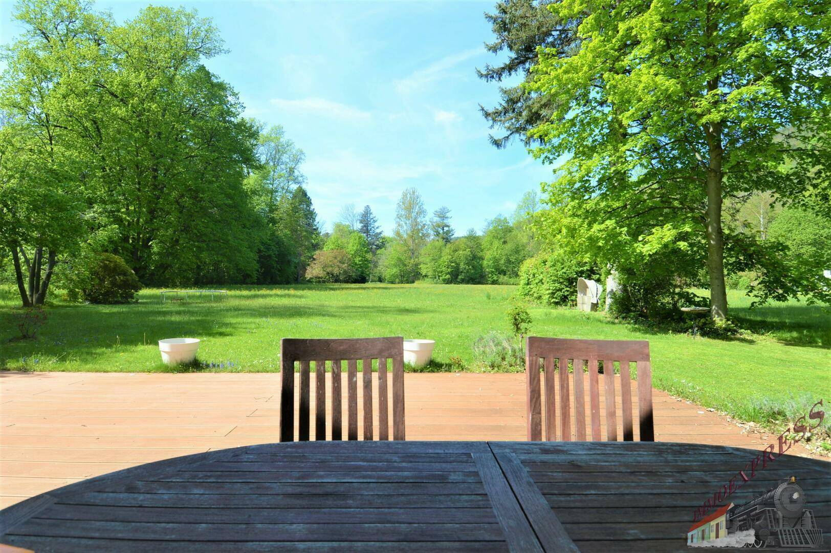 Holzterrasse mit Gartenmöbeln und weitem Blick über eine grüne Wiese und Bäume.