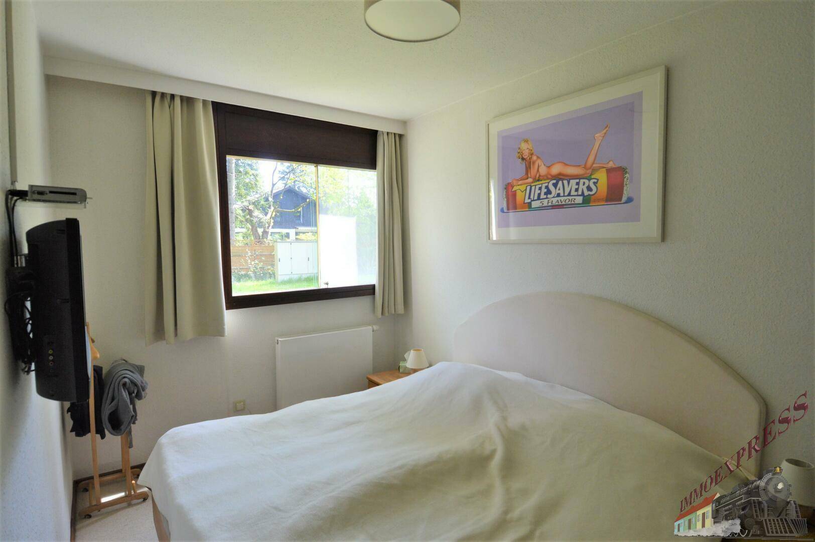 Helles Schlafzimmer mit Doppelbett, Fenster und einem an der Wand montierten Fernseher.