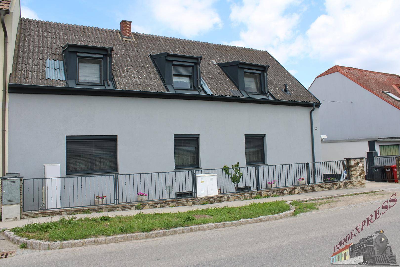 Vorderansicht des grauen Hauses mit dunklen Fenstern, Gauben und einem gepflegten Vorgartenbereich.