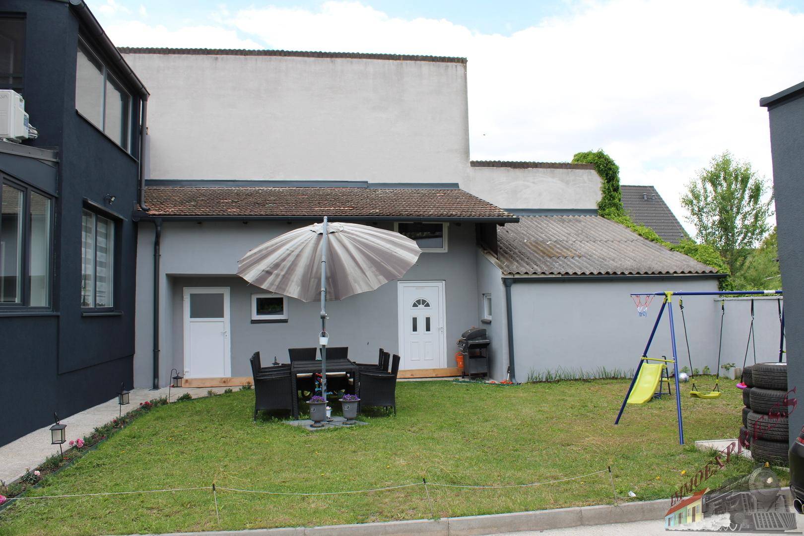 Hinterhof mit Rasenfläche, Gartenmöbeln unter einem Sonnenschirm und einem Kinderspielplatz.