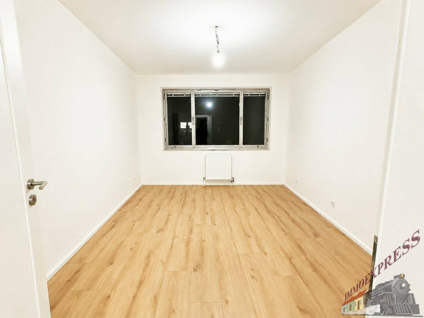 Unmöbliertes Zimmer mit hellem Laminatboden und einem großen Fenster, das viel Licht hereinlässt.