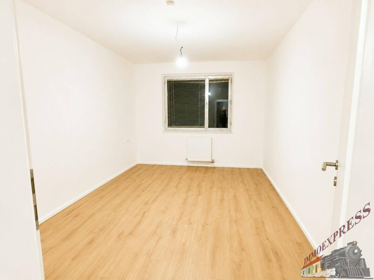 Leeres Zimmer mit Laminatboden, weißen Wänden und einem Fenster mit Jalousien.