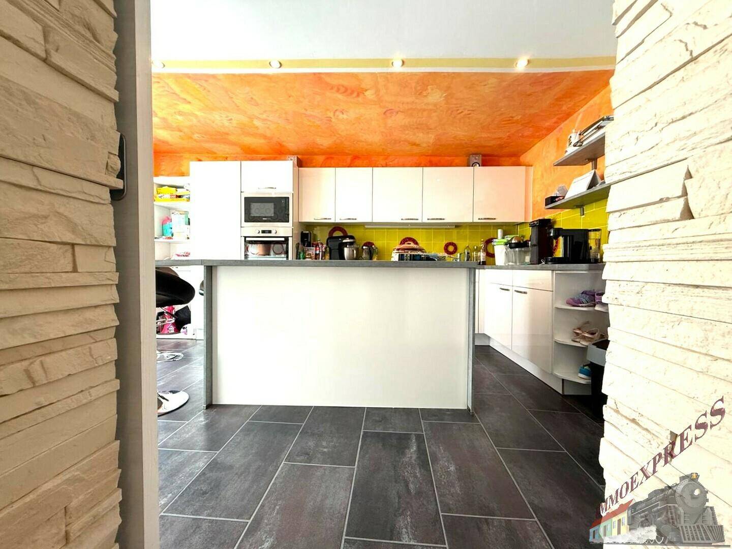 Blick vom Flur in die moderne Küche mit weißen Schränken, Kücheninsel und orangefarbener Decke.