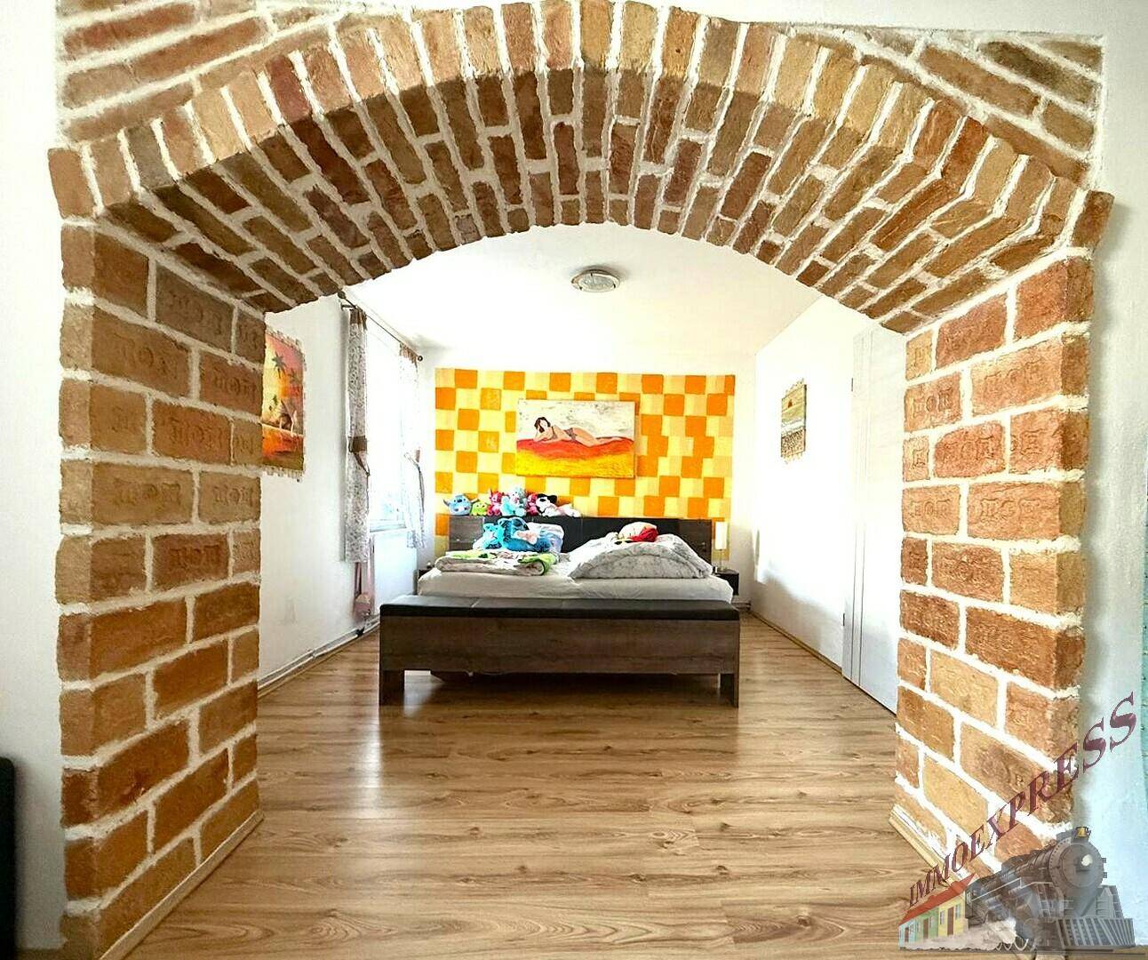 Blick durch den Ziegelbogen in den Schlafbereich mit einem Bett und einer farbenfrohen Wandgestaltung.