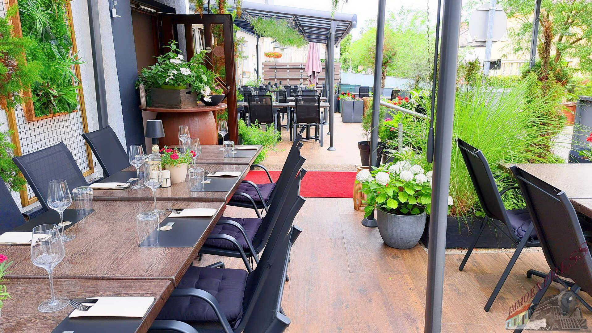 Helle Restaurantterrasse mit Holzboden, modernen Tischen und Stühlen, umgeben von Grünpflanzen.