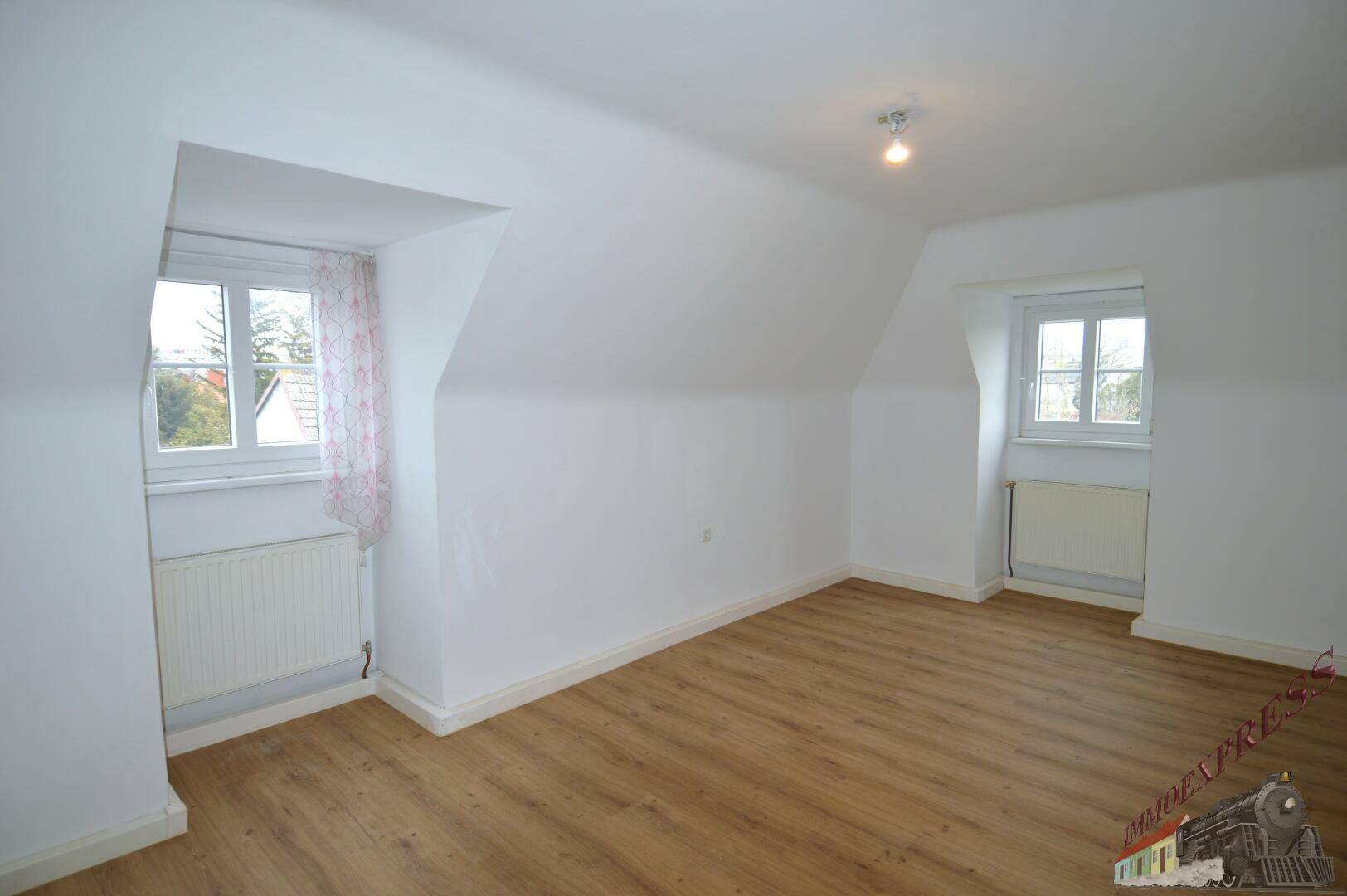 Helles Dachzimmer mit schrägen Wänden, Laminatboden und zwei Fenstern.