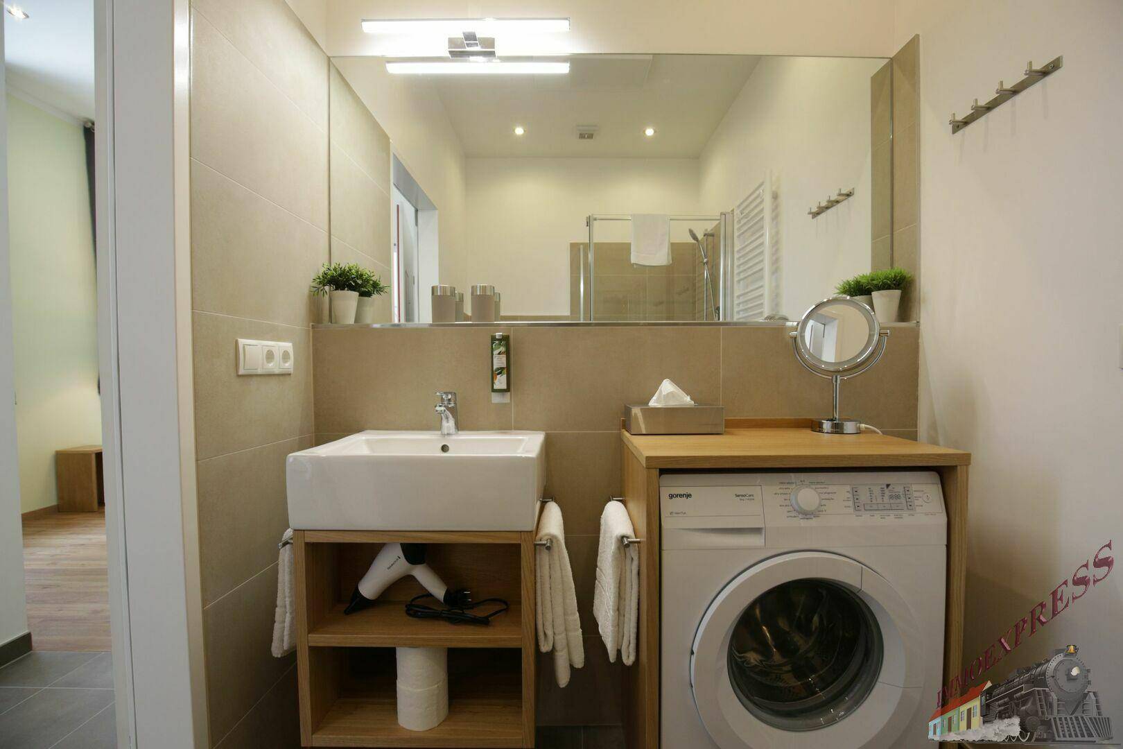 Modernes Badezimmer mit Waschbecken, Waschmaschine, großem Spiegel und beigefarbenen Fliesen.