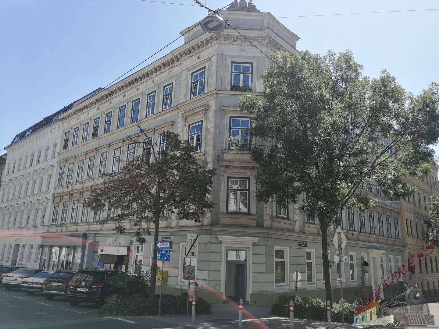 Eckgebäude mit klassischer Architektur, vielen Fenstern und Bäumen an einer städtischen Straße.