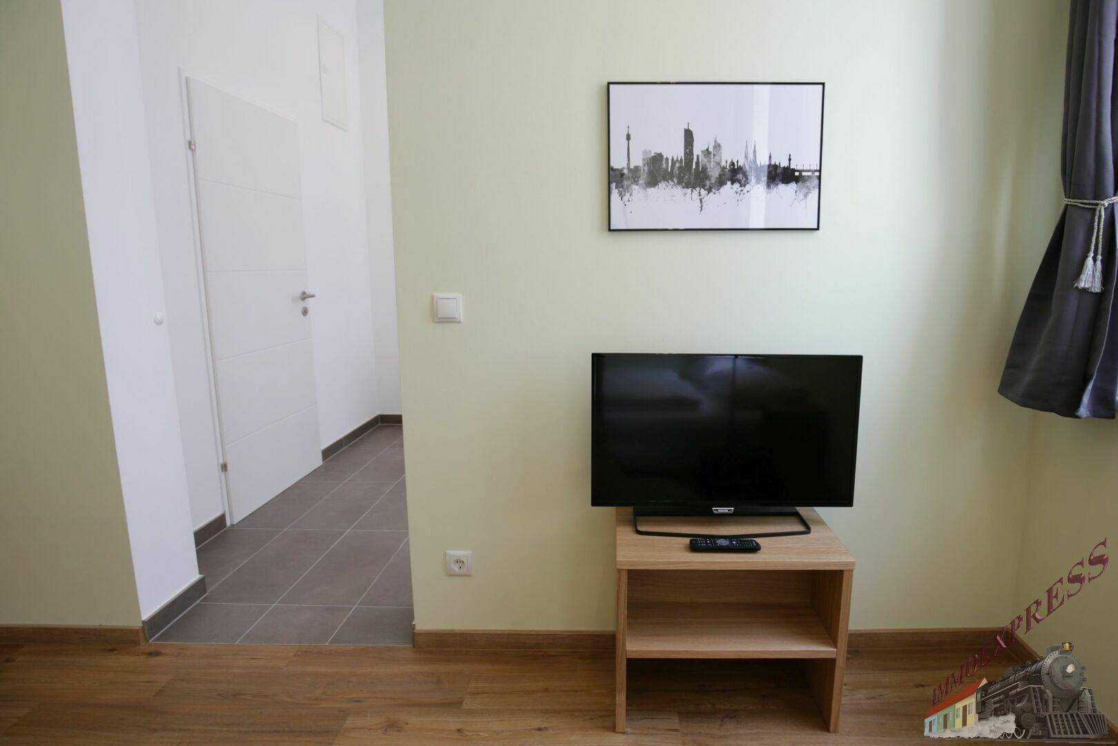 Heller Wohnbereich mit Holzboden, einem Fernseher auf einem kleinen Tisch und einem Flur im Hintergrund.