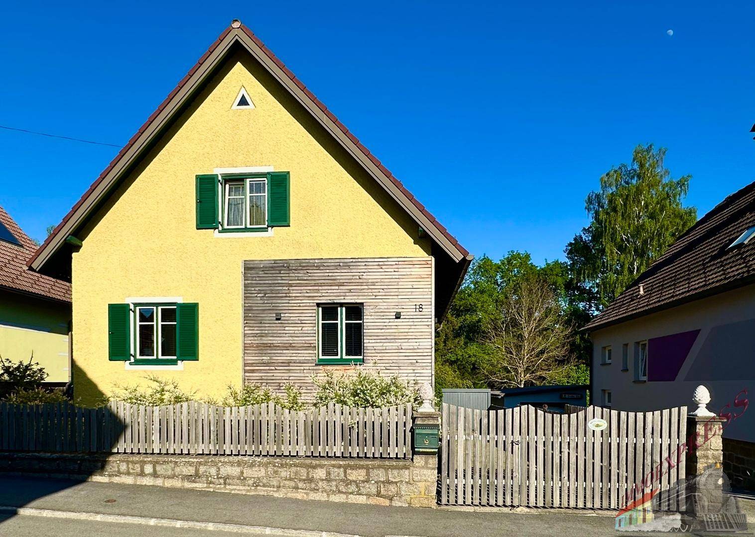 Gelbes Einfamilienhaus mit grünen Fensterläden und Holzelementen unter blauem Himmel.